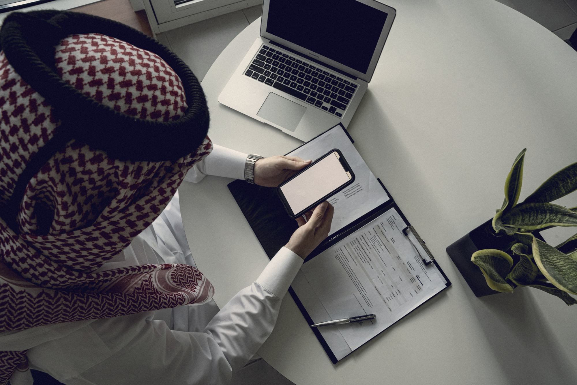 A Qatari professional reviewing a strategy document with phone and laptop